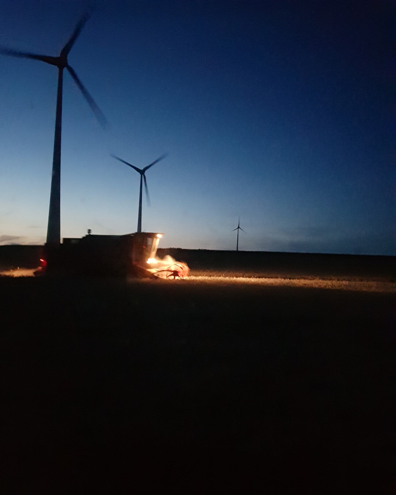 Foto: Eine nächtliche Szene zeigt mehrere Windkraftanlagen auf einem Feld. Im Vordergrund ist ein beleuchtetes landwirtschaftliches Fahrzeug bei der Arbeit zu sehen.