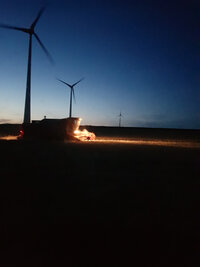 Foto: Eine nächtliche Szene zeigt mehrere Windkraftanlagen auf einem Feld. Im Vordergrund ist ein beleuchtetes landwirtschaftliches Fahrzeug bei der Arbeit zu sehen.