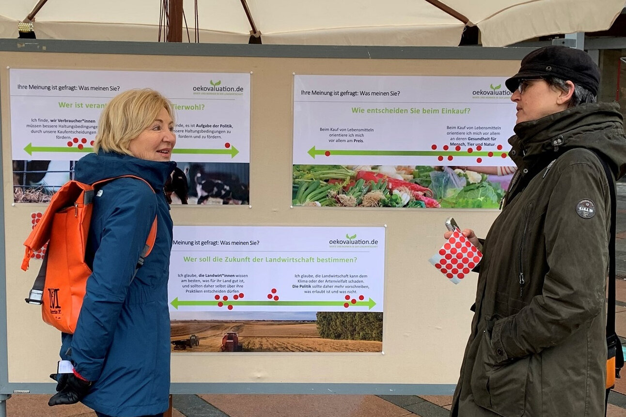Foto: Zwei Personen stehen vor Plakaten mit Fragen zur Landwirtschaft und Tierwohl. Die Plakate tragen die Überschrift 'Ihre Meinung ist gefragt: Was meinen Sie?' und zeigen Themen wie Tierwohl und Einkaufskriterien.