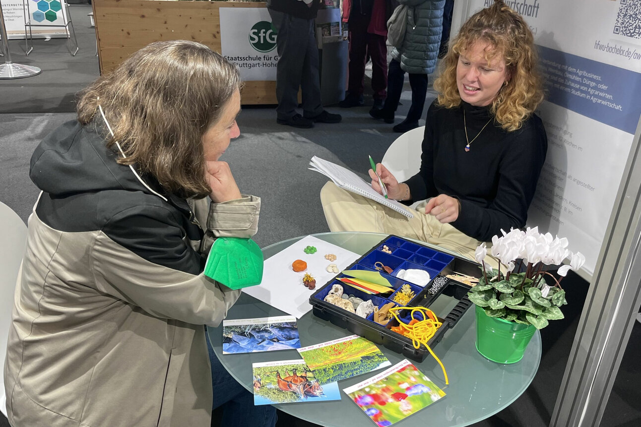 Foto: Zwei Personen sitzen an einem Tisch mit Pflanzen und Materialien. Eine Person schreibt in ein Notizbuch, während die andere zuhört. Im Hintergrund sind Messewände und ein Schild mit der Aufschrift 'Staatsschule für Gartenbau Stuttgart-Hohenheim' sichtbar.