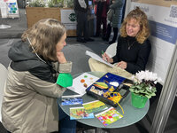 Foto: Zwei Personen sitzen an einem Tisch mit Pflanzen und Materialien. Eine Person schreibt in ein Notizbuch, während die andere zuhört. Im Hintergrund sind Messewände und ein Schild mit der Aufschrift 'Staatsschule für Gartenbau Stuttgart-Hohenheim' sichtbar.