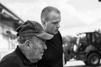 Foto: Zwei Personen stehen im Freien. Eine Person trägt eine Mütze. Im Hintergrund ist ein unscharfes Fahrzeug zu sehen.