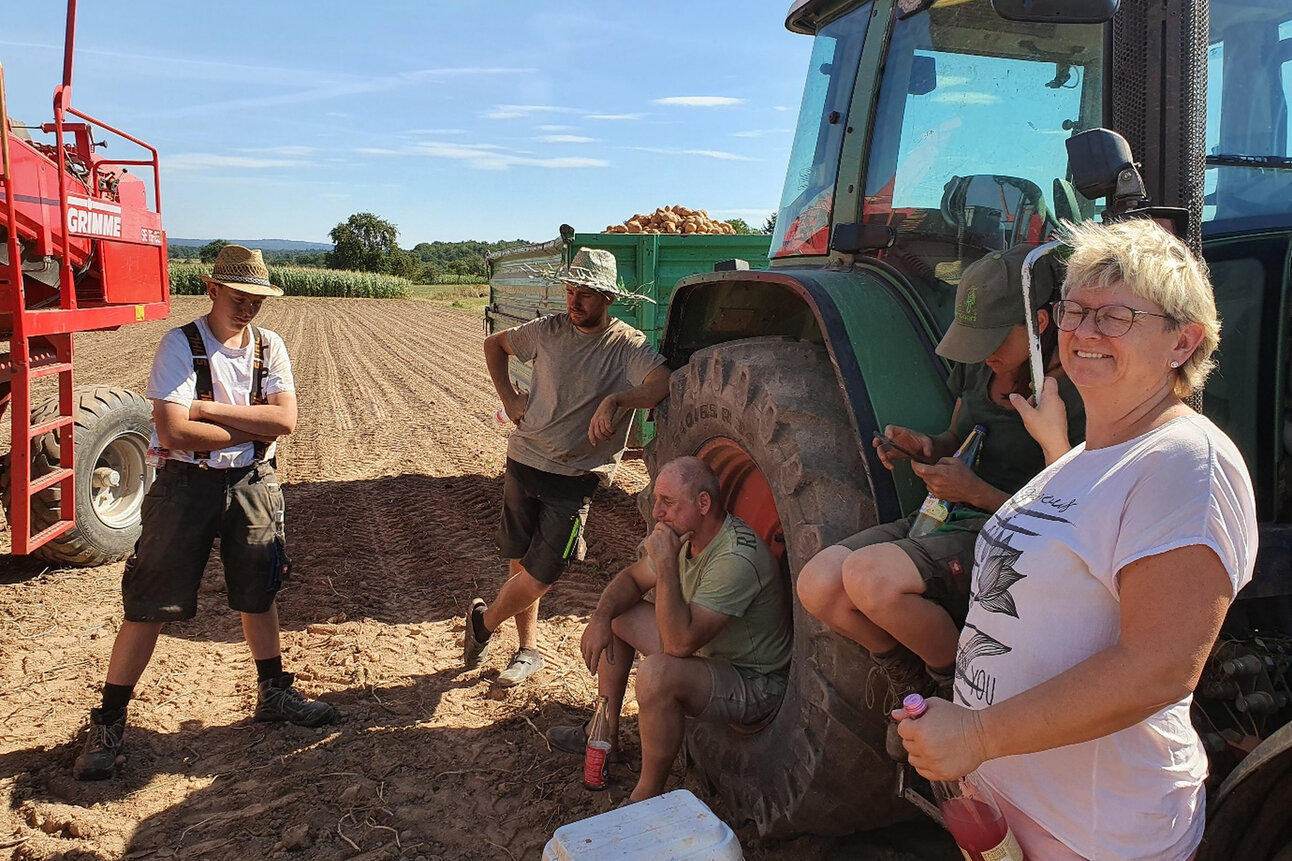 Foto: Mehrere Personen machen eine Pause neben einem Traktor auf einem Feld. Eine Person hält eine Flasche, während andere sitzen oder stehen. Im Hintergrund ist landwirtschaftliches Gerät und ein gepflügtes Feld zu sehen.
