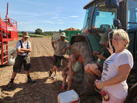 Foto: Mehrere Personen machen eine Pause neben einem Traktor auf einem Feld. Eine Person hält eine Flasche, während andere sitzen oder stehen. Im Hintergrund ist landwirtschaftliches Gerät und ein gepflügtes Feld zu sehen.