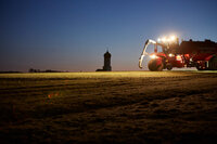 Foto: Ein Traktor mit Beleuchtung arbeitet auf einem Feld bei Sonnenuntergang. Im Hintergrund ist die Silhouette einer Kirche mit Zwiebelturm sichtbar.
