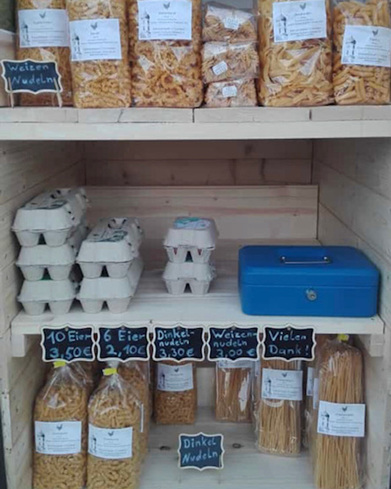 Foto: Ein Regal mit verschiedenen Nudelsorten und Eierkartons. Oben sind Weizennudeln, unten Dinkelnudeln und Eier. Preisschilder zeigen 10 Eier für 3,50€, 6 Eier für 2,10€, Dinkelnudeln für 3,30€ und Weizennudeln für 3,00€.