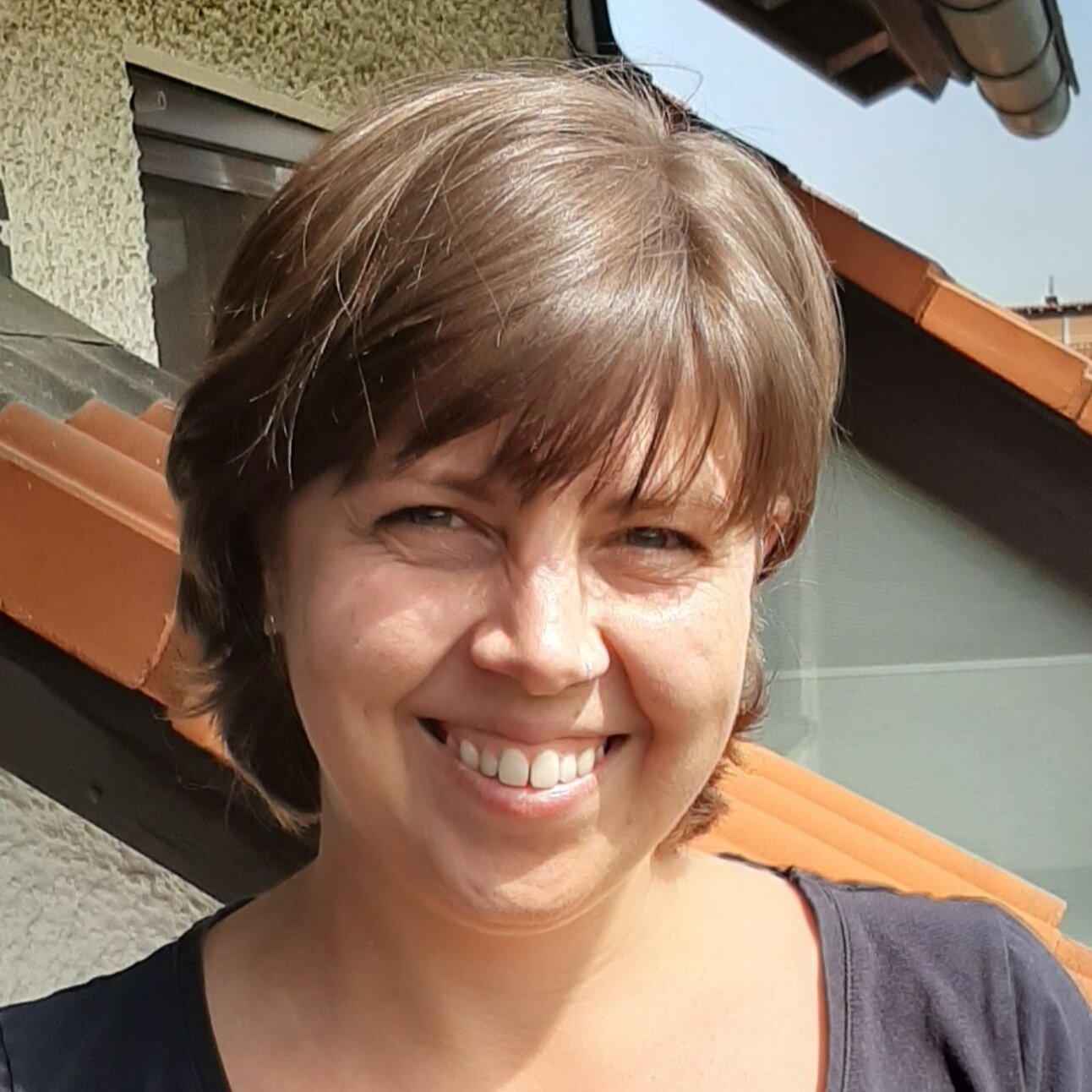 Foto: Eine Person steht auf einem Balkon mit Dachziegeln im Hintergrund. Die Umgebung zeigt ein Gebäude mit verputzter Fassade und einem Fenster.