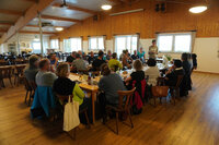 Foto: Eine Gruppe von Personen sitzt in einem Raum mit Holzwänden und -boden an einem großen Tisch. Eine Person steht und spricht zu den anderen.