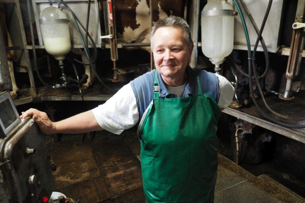 Foto: Eine Person in einem grünen Schürze steht in einer Molkerei. Im Hintergrund sind Milchtanks und Schläuche sichtbar. Die Person hält sich an einem Steuerpult fest.