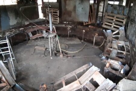 Foto: Eine verlassene industrielle Umgebung mit Metallstrukturen, Paletten und einer Leiter. Der Raum wirkt ungepflegt und enthält verschiedene Metallteile und Kabel.