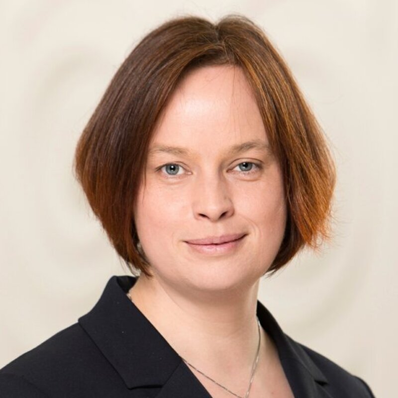 Foto: Claudia Bieling trägt einen dunklen Blazer und eine Kette vor einem neutralen Hintergrund.