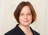 Foto: Claudia Bieling trägt einen dunklen Blazer und eine Kette vor einem neutralen Hintergrund.