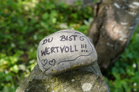 Foto: Ein Stein liegt auf einem anderen Stein in einer natürlichen Umgebung. Auf dem Stein steht in schwarzer Schrift 'DU BIST WERTVOLL !!!' mit zwei gezeichneten Herzen.