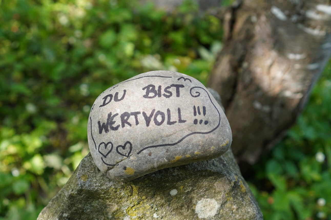 Foto: Ein Stein liegt auf einem anderen Stein in einer natürlichen Umgebung. Auf dem Stein steht in schwarzer Schrift 'DU BIST WERTVOLL !!!' mit zwei gezeichneten Herzen.