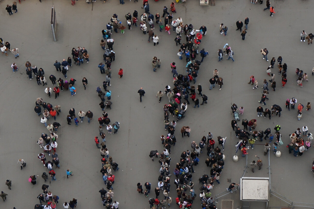 Foto: Eine große Gruppe von Menschen steht in mehreren Schlangen auf einem offenen Platz. Die Personen sind aus der Vogelperspektive zu sehen und tragen verschiedene Kleidung.