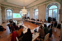Foto: Personen sitzen in einem historischen Raum mit Kronleuchter an langen Tischen. Eine Präsentation wird auf eine Leinwand projiziert. Große Fenster lassen Tageslicht herein.