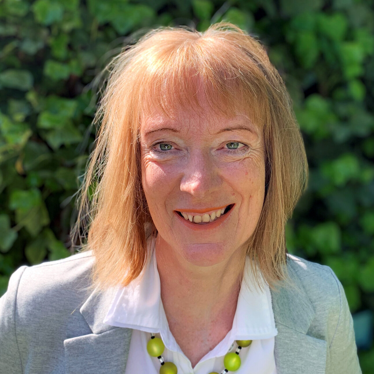 Foto: Jutta Schneider-Rapp in einem grauen Blazer und weißem Hemd steht vor einem Hintergrund aus grünen Blättern. Die Person trägt eine Halskette mit großen grünen Perlen.