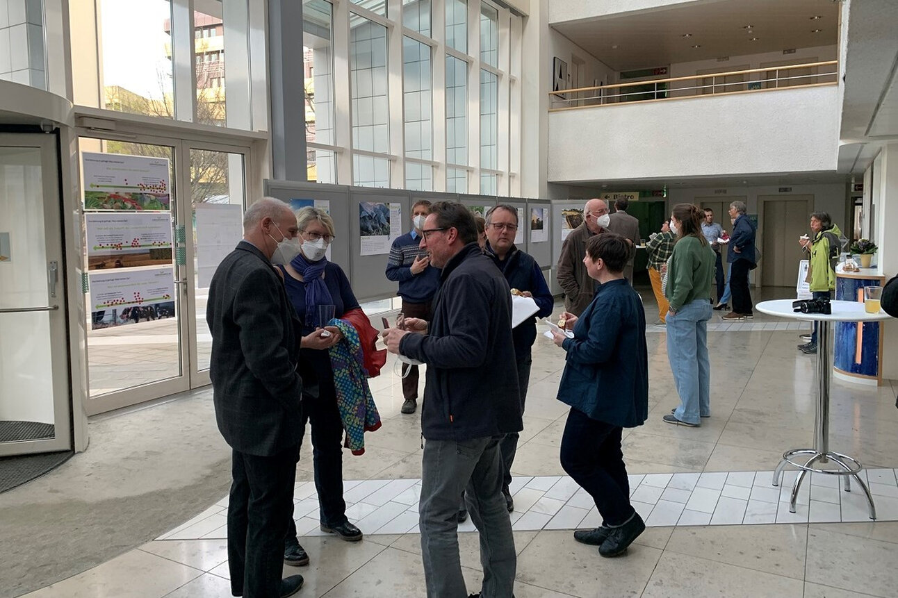 Foto: Eine Gruppe von Personen steht in einem modernen Foyer mit großen Fenstern. Im Hintergrund sind Ausstellungstafeln und weitere Personen sichtbar.
