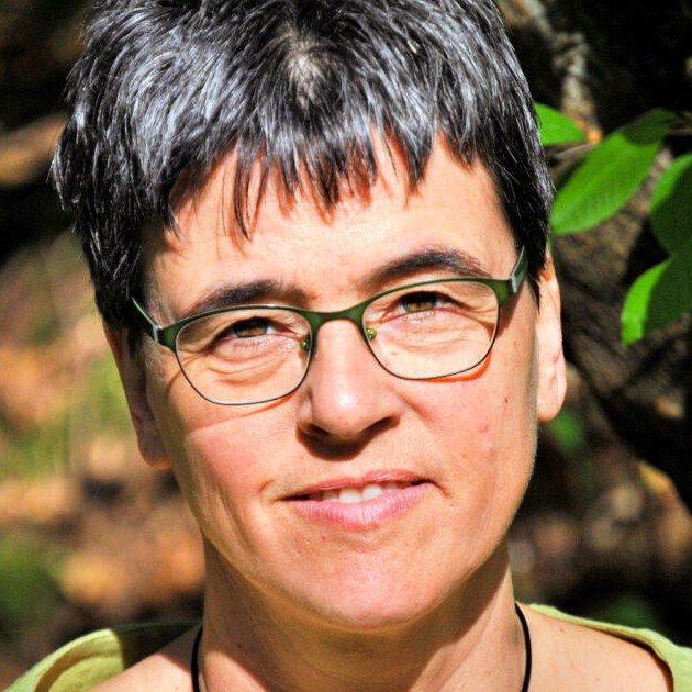 Foto: Uta Eser steht im Freien, umgeben von grünen Blättern. Sonnenlicht fällt auf die Szene, was auf eine natürliche Umgebung hindeutet.
